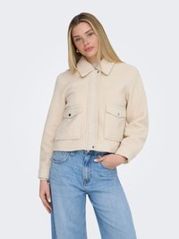 Kvinde med langt blondt hår if ørt en beige cropped uldjakke med forlommer og lyseblå højtaljede jeans mod en hvid baggrund.