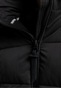 Czarna kurtka puchowa z grubą, pikowaną teksturą, wysokim kołnierzem i wyraźnym zamkiem z logo "SUPERDRY."