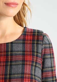 Haut en plaid en laine avec des motifs rouges, verts et jaunes sur un fond gris ; col arrondi et manches bouffantes pour un détail de design.