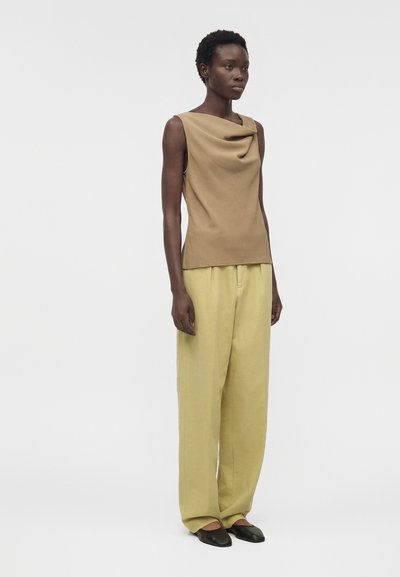 Γυναίκα στέκεται φορώντας ένα αμάνικο καφέ τοπ με ντραπέ ντεκολτέ, άνετο κίτρινο παντελόνι και μαύρα επίπεδα παπούτσια σε απλό φόντο.