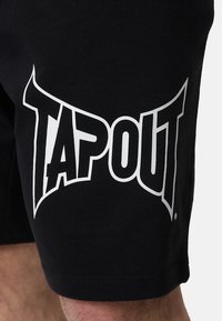 Crne kratke hlače od pamuka, s velikim bijelim logotipom "TAPOUT" otisnutim na lijevoj nozi. Tkanina izgleda glatko s klasičnim krojem.