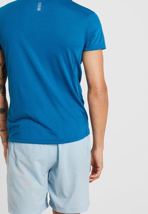 Personne portant une chemise à manches courtes bleue et un short de sport bleu clair, vue de dos sur un fond uni.