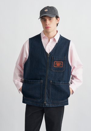 Fiatal férfi fekete sapkában, rózsaszín gombos ingben és sötétkék farmer mellényben, elülső zsebekkel és narancssárga Dickies logóval.