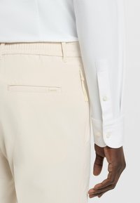 Vit långärmad skjorta med texturerat tyg och knappmanschetter, ihop med beige byxor med zip-ficka och diskret logotyp.