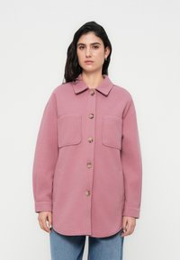 Ροζ, oversized σακάκι με κουμπιά, κατασκευασμένο από μαλακό ύφασμα. Διαθέτει δύο μπροστινές τσέπες και κλασικό γιακά. Ελκυστική υφή με ορατές ραφές.