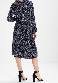 Abito blu navy con pois bianchi, maniche lunghe e schiena arricciata, stretto in vita. Indossato con sandali neri con tacco alto.