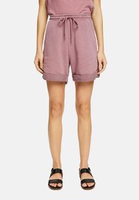 Femme portant un short à cordon de serrage mauve et des sandales noires ouvertes à l'avant, debout contre un fond blanc uni.