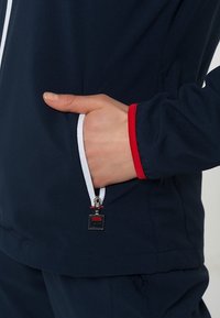 Marinblå idrottsjacka med dragkedjeficka, med röda och vita detaljer vid ärmslut och fickans dragkedja, slät tygkvalitet.