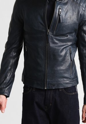 Chaqueta de cuero negro con cremallera frontal y tres bolsillos con cremallera, usada sobre vaqueros oscuros por una persona con los brazos relajados a los lados.