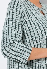 Distanza ravvicinata del braccio e della parte superiore del torso di una persona che indossa una camicia a fantasia con base verde e forme rettangolari arrotondate chiare.
