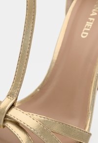 Lähikuva kultaisista metallinhohtoisista strappy-korkokengistä, joissa on beige sisäpohja ja "Via Field" -brändin logo näkyvissä.