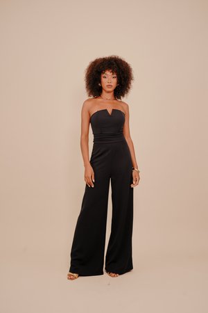Woman with curly hair stands wearing a strapless black jumpsuit and gold sandals against a beige background.