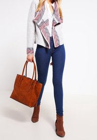 Hnědá kožená kabelka s dvojitými uchy, nošená s světle šedým kardiganem s ozdobným límečkem, ve spojení s tmavě modrými úzkými džínami a hnědými kotníkovými botami.