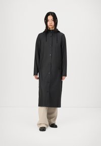 Lång svart regnkappa med huva, tryckknappar och sidofickor, tillverkad av slät, vattenavvisande tyg. Bärs över beige byxor.
