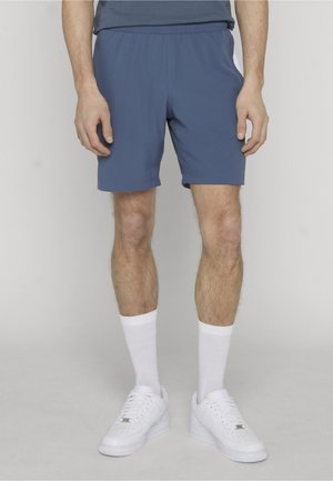 Man die blauwe sportkorte broek, witte sportsokken en witte sneakers draagt, staat met ontspannen handen langs zijn zij tegen een neutrale achtergrond.