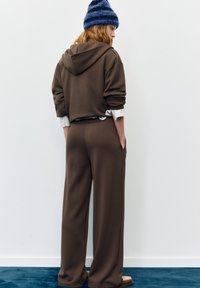 Brown hoodie and wide-leg pants made of soft material. Model wears a blue striped beanie. Simple design with functional pockets.