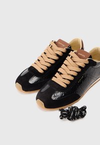 Czarne sneakersy z połączeniem zamszu i błyszczącej skóry lakierowanej, z beżowymi sznurówkami, brązową naszkliwo z logo "NUBIKK" i gumową podeszwą.