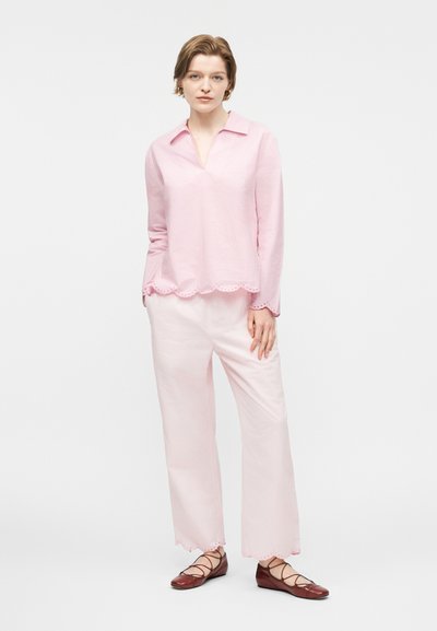 Nő világos rózsaszín hosszú ujjú blúzban és hozzá illő, fodros szélű bő szárú nadrágban, barna fűzős lapos cipővel, áll.