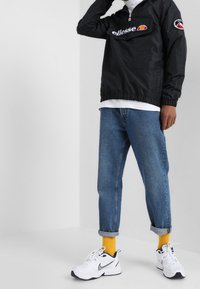 Sort vindjakke med logo, grå T-shirt, falmede blå jeans og hvide sneakers. Omslåede ærmer og gule sokker tilføjer kontrast.
