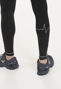 Zwarte leggings met een witte hartslagpatroon nabij de kuiten, gecombineerd met donkere hardloopschoenen met een mesh bovenkant en een gevoerde zool.