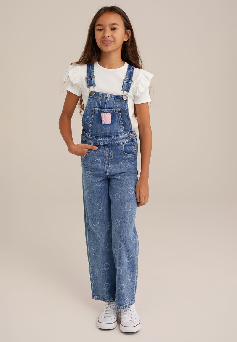 Pantalones de mezclilla con un corte relajado, de color azul claro con un patrón circular, tirantes ajustables y un parche de bolsillo rosa. Combinados con una blusa blanca con volantes.