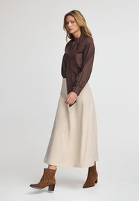 Kvinde, der går i en beige lang nederdel, brun bluse med bindebånd og brune ruskinds ankelstøvler mod en ensfarvet hvid baggrund.