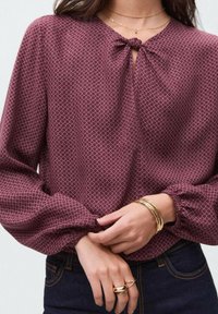 Bordeauxfarbige Bluse mit geometrischem Muster, Schleifenkragen und Puffärmeln. Verfügt über elastische Bündchen und geschichtete metallische Armbänder am Handgelenk.