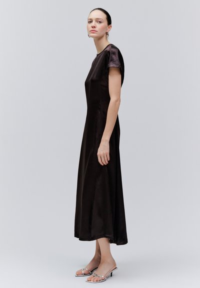 Femme debout portant une robe midi en satin noire à manches courtes et des escarpins argentés à bout ouvert, sur un fond gris clair uni.