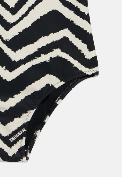 Tissu à motif zigzag noir et blanc avec des coutures visibles et un petit logo texte blanc dans le coin inférieur gauche.