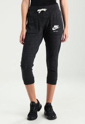 Persona lleva pantalones deportivos negros recortados con el logo de Nike y cordón blanco, combinados con zapatillas deportivas negras y una camiseta negra.