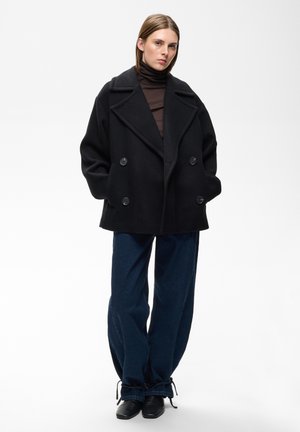 Femme portant un manteau noir oversize à double boutonnage, un col roulé sombre, un jean bleu foncé ample avec des liens aux ourlets, et des chaussures noires.