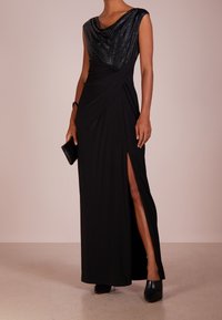 Svart maxiklänning med sjalkrage, glittrigt överstycke, sidospalt och draperad design. Bärs med svart clutch och högklackade skor.