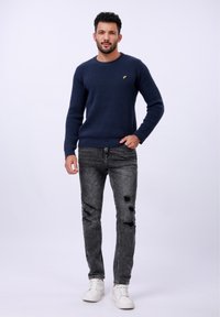 Pull marine texturé, jeans noirs déchirés et baskets blanches. Le pull arbore un petit logo jaune. Tenue décontractée avec une coupe ample.