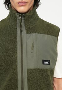 Olijfgroen vest met een mix van zachte fleece en nylon, hoge kraag, ritssluiting aan de voorkant en een label aan de onderzijde rechts.