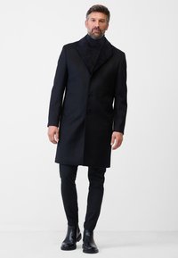 Schwarzer Wollmischmantel mit einem strukturierten, marineblauen Kragen, einreihigem Design, fünf Knöpfen, kombiniert mit schwarzen Hosen und Stiefeletten.