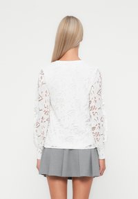 Blusa bianca con intricate maniche lunghe in pizzo, collo rotondo e schiena liscia. Abbinata a una gonna grigia plissettata, che mostra una texture a strati.