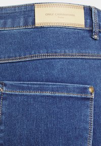 Jeans in denim blu con un'etichetta marrone chiaro contrassegnata "ONLY CARMAKOMA". Presentano rivetti in rame e cuciture a contrasto sulla tasca posteriore.