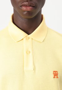 Žltý polo tričko vyrobené z textúrovanej látky, s trojicou gombíkov na výstrihu a s oranžovým logom na ľavej hrudi.