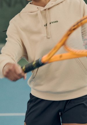 Person i beige hættetrøje og sorte shorts holder et orange tennis ketsjer og gør sig klar til at slå en bold på en indendørs bane.
