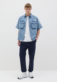 Camisa de mezclilla de manga corta en azul claro con dos bolsillos frontales, llevada sobre una camiseta blanca. Combinada con pantalones cargo azul marino y zapatillas blancas.