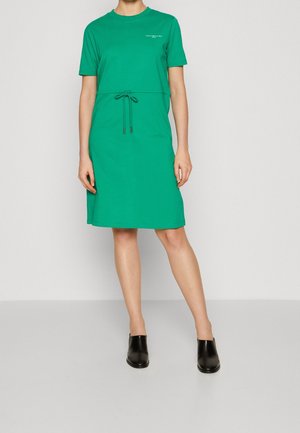 Women’s green knee-length dress with short sleeves, drawstring waist, small chest logo, paired with black slip-on shoes.