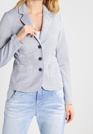 Femme portant un blazer gris clair texturé à trois boutons et poches plaquées, associé à un jean en denim délavé clair.