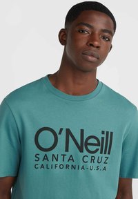 Tyrkysové tričko s krátkým rukávem a kulatým výstřihem. Na přední straně se nachází tučný černý text: "O'Neill SANTA CRUZ CALIFORNIA-U.S.A."