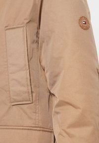Tan pufferjack met een zijzak, gestructureerde stof en een cirkelvormig logo in de kleuren rood, wit en blauw op de schouder.