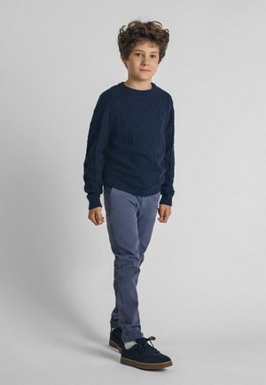 Granatowy sweter w splot warkoczowy, jasnoniebieskie spodnie slim-fit i ciemne sneakersy. Gładki przód, elastyczne mankiety i teksturowany wzór plecionki na swetrze.