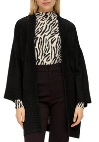 Schwarze Strickjacke mit offenem Vorderteil, weiten Ärmeln und gerippter Textur. Getragen über einem hochgeschlossenen, schwarz-weiß gestreiften Oberteil.