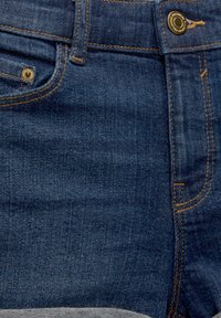 Tmavě modré džínové kalhoty s jemnou texturou, vybavené kovovými doplňky v zlaté barvě a kontrastním světle hnědým stehem podél kapes a švů.