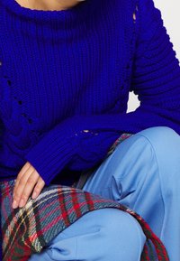 Personne portant un pull en laine bleu vif et un pantalon bleu clair, posant la main sur un tissu à carreaux rouge et vert.