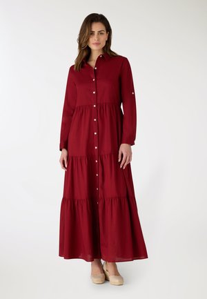 Donna che indossa un abito maxi rosso a maniche lunghe, con bottoni e balze, abbinato a scarpe beige con zeppa, in piedi davanti a uno sfondo bianco semplice.