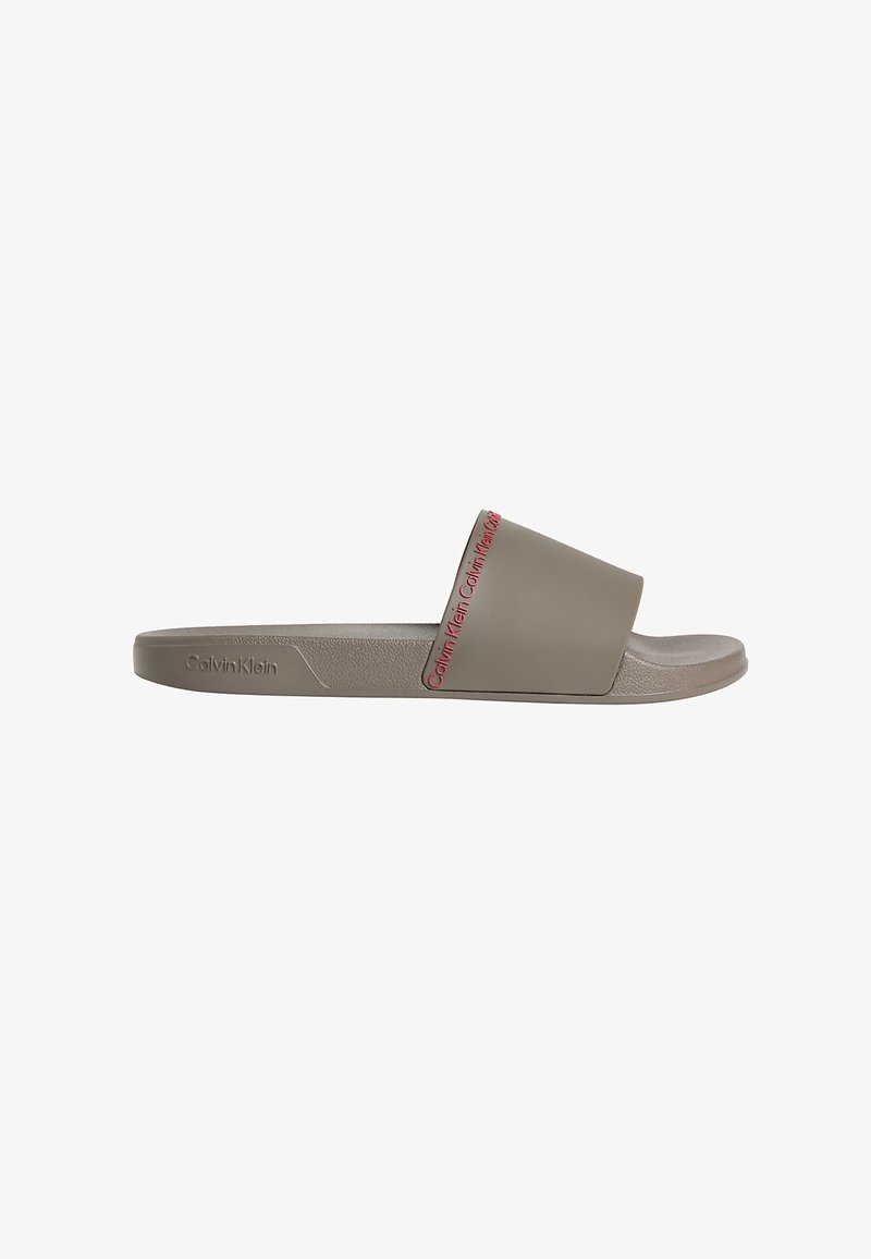 Sandale mule taupe unique avec une large bride portant le texte du logo Calvin Klein en rouge, semelle texturée et logo embossé sur le côté.